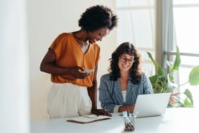 black woman holding iphone talking to white woman working on laptop
